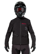 Pilote de motocross portant la veste softshell Alpinestars Lite-Dura noire, avec des détails de logos rouge et gris sur la poitrine, casque noir avec lunettes teintées en rouge, gants et pantalon noirs, vue de face d'un équipement de conduite technique tout-terrain