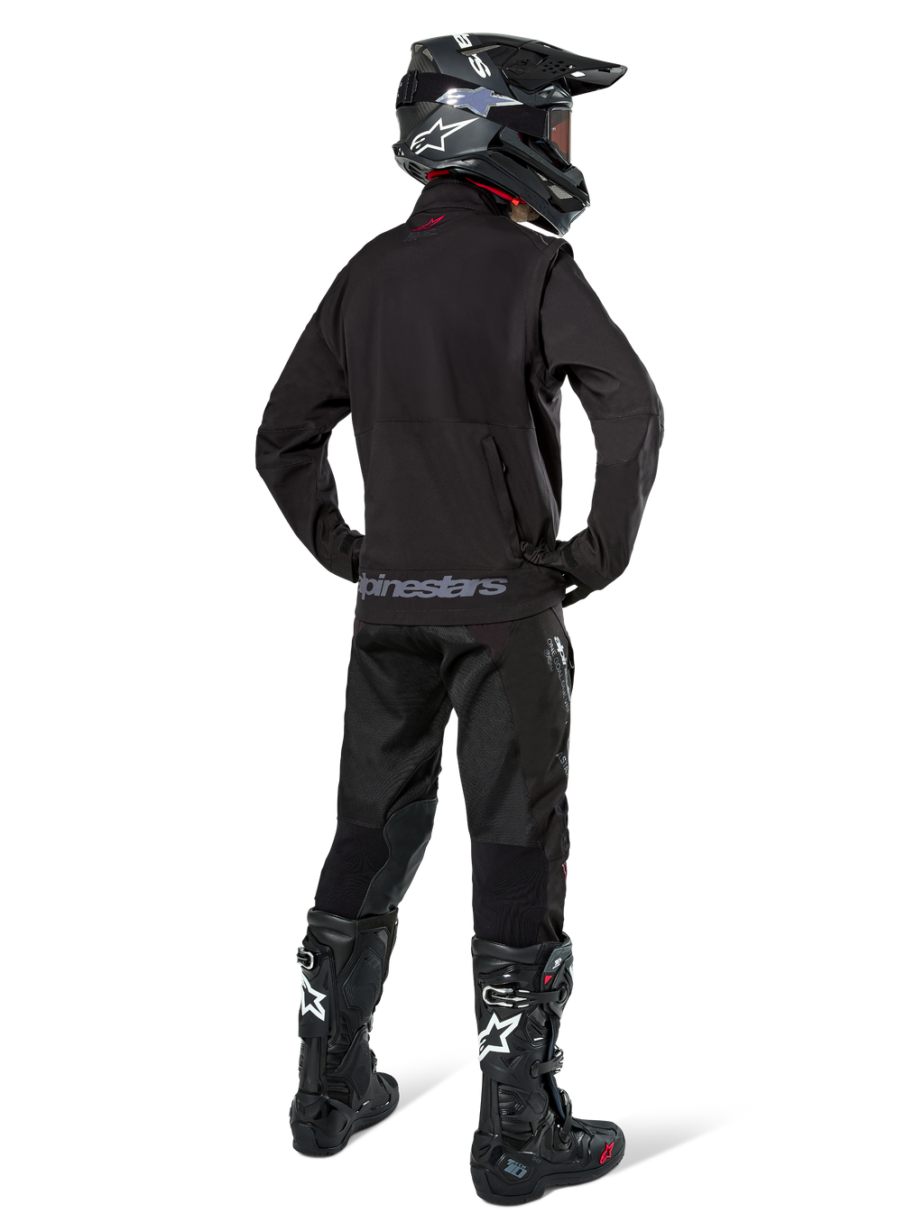 Pilote d'enduro vu de dos portant la veste softshell Alpinestars Lite-Dura en noir, associée à un pantalon technique et des bottes noirs, avec poche de rangement arrière zippée et logo Alpinestars sur l'ourlet inférieur, équipement tout-terrain complet pour le motocross.
