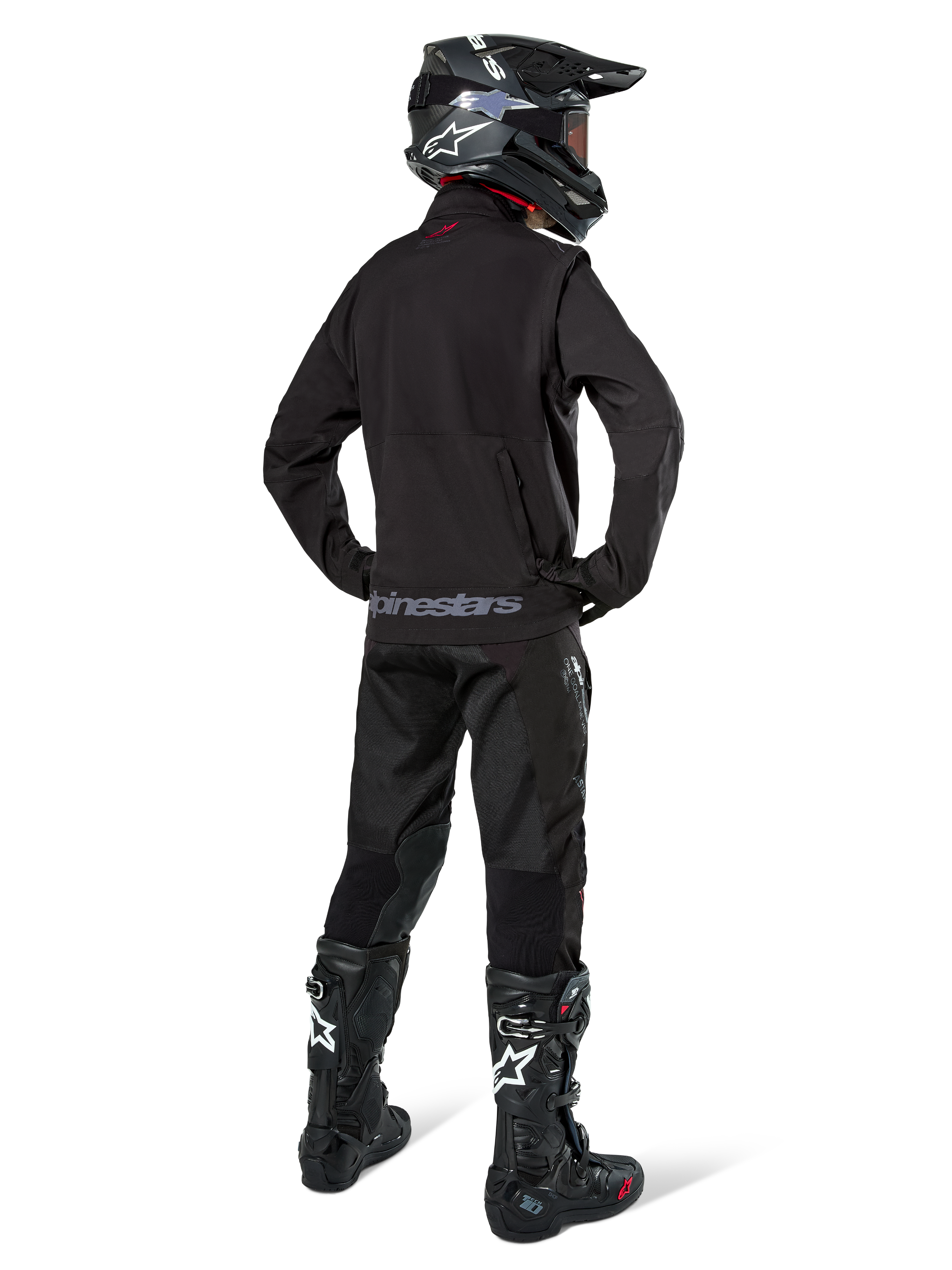 Pilote d'enduro vu de dos portant la veste softshell Alpinestars Lite-Dura en noir, associée à un pantalon technique et des bottes noirs, avec poche de rangement arrière zippée et logo Alpinestars sur l'ourlet inférieur, équipement tout-terrain complet pour le motocross.