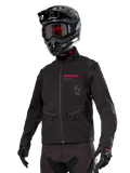 Alpinestars Lite-Dura Softshell Jacket, veste de moto tout-terrain noire avec logos rouges et logo AS, portée avec un casque de motocross noir, des lunettes rouges et des gants noirs, dotée de poches zippées et de poignets réglables, conçue pour l'enduro