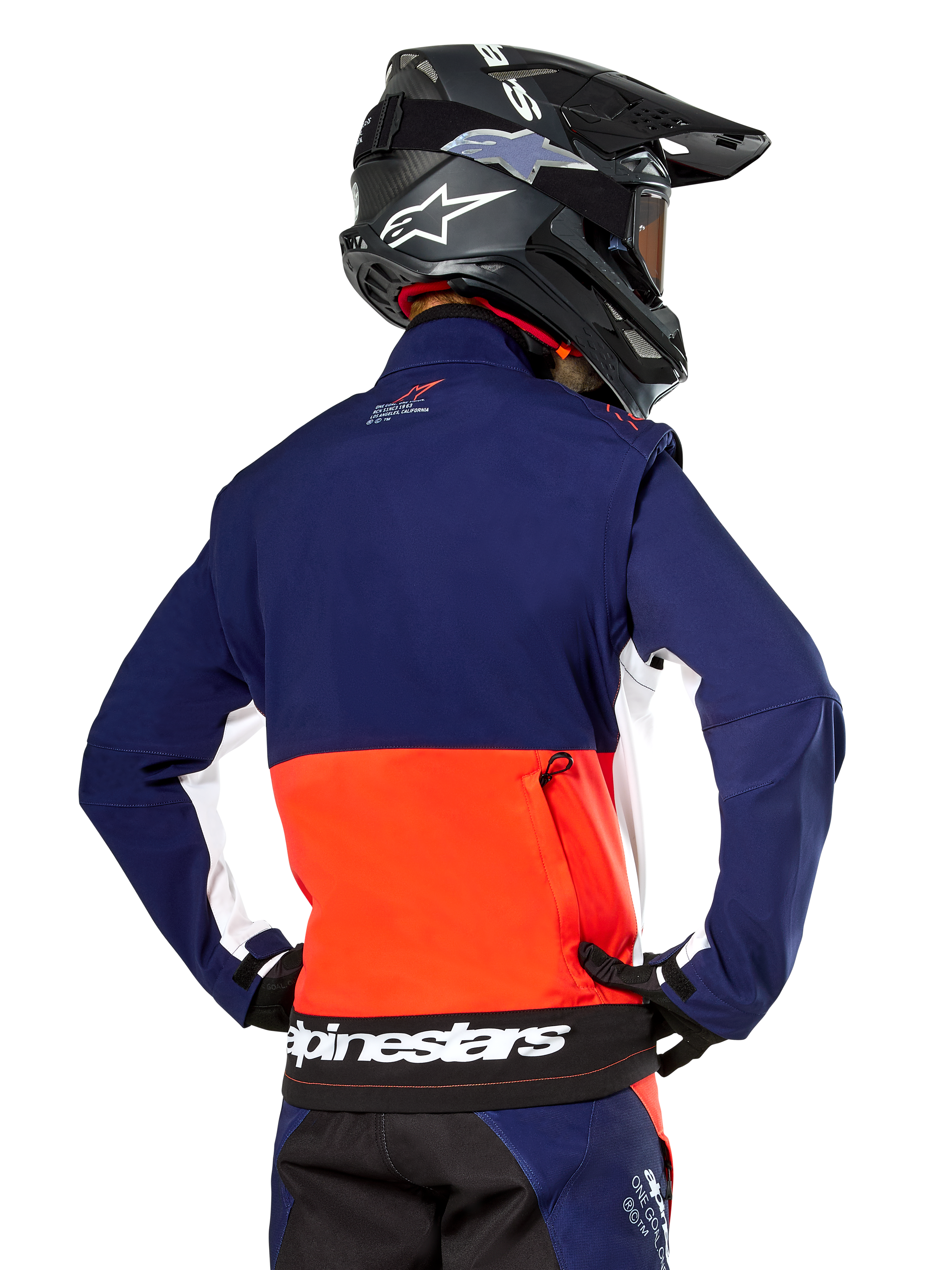 Veste Softshell Alpinestars Lite-Dura, bleu marine, orange vif et blanc, vue arrière portée par un pilote de motocross, avec un grand logo blanc sur l'ourlet arrière et une poche de rangement zippée, faisant partie d'une tenue de course tout-terrain complète.