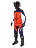 Pilote de motocross portant la veste Softshell Alpinestars Lite-Dura en Navy, Orange Vif et Blanc, avec pantalon tout-terrain assorti, casque noir avec lunettes et bottes de course noires, tenue d'enduro coordonnée