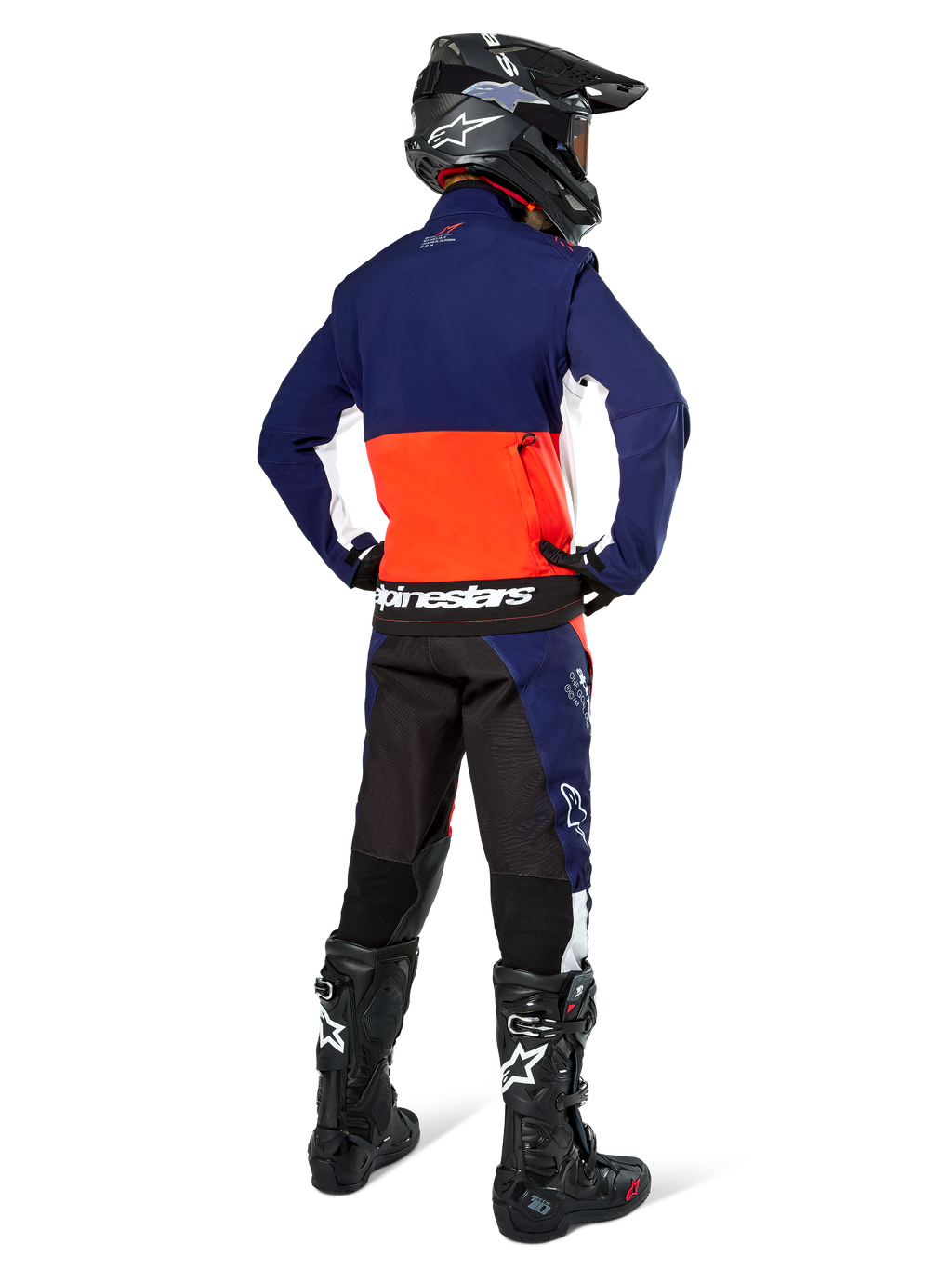 Pilote de motocross vu de dos portant la veste Softshell Alpinestars Lite-Dura en bleu marine, orange vif et blanc, montrant la grande poche de rangement arrière, avec un pantalon d'enduro assorti, un casque de motocross noir et des bottes tout-terrain noires