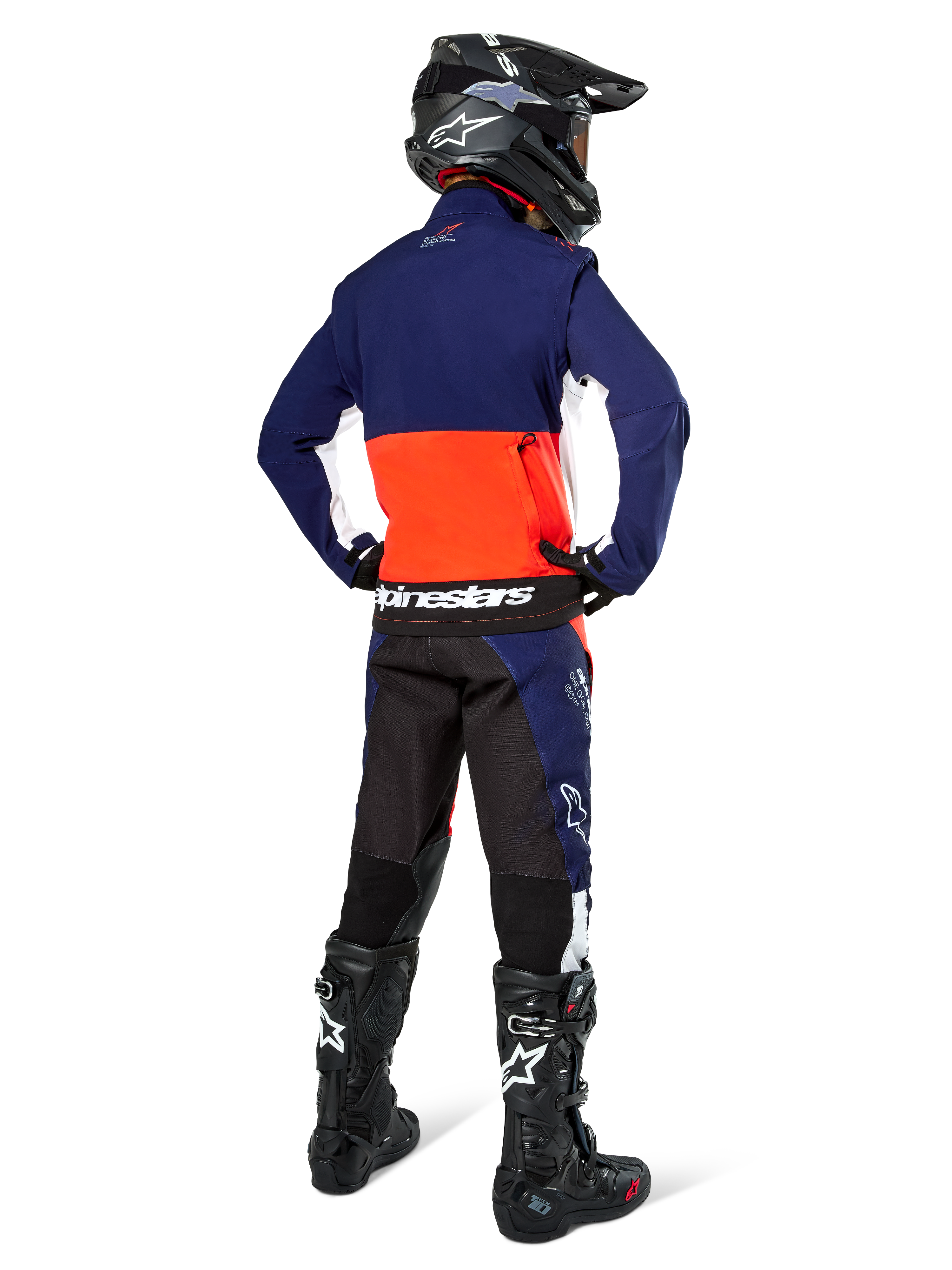 Pilote de motocross vu de dos portant la veste Softshell Alpinestars Lite-Dura en bleu marine, orange vif et blanc, montrant la grande poche de rangement arrière, avec un pantalon d'enduro assorti, un casque de motocross noir et des bottes tout-terrain noires