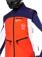 Veste Softshell Alpinestars Lite-Dura en bleu marine, orange vif et blanc, vue rapprochée du torse, empiècement frontal orange vif, panneaux latéraux blancs et épaules bleu marine, poches zippées, logo Alpinestars bien visible, portée par un pilote avec un casque tout-terrain noir