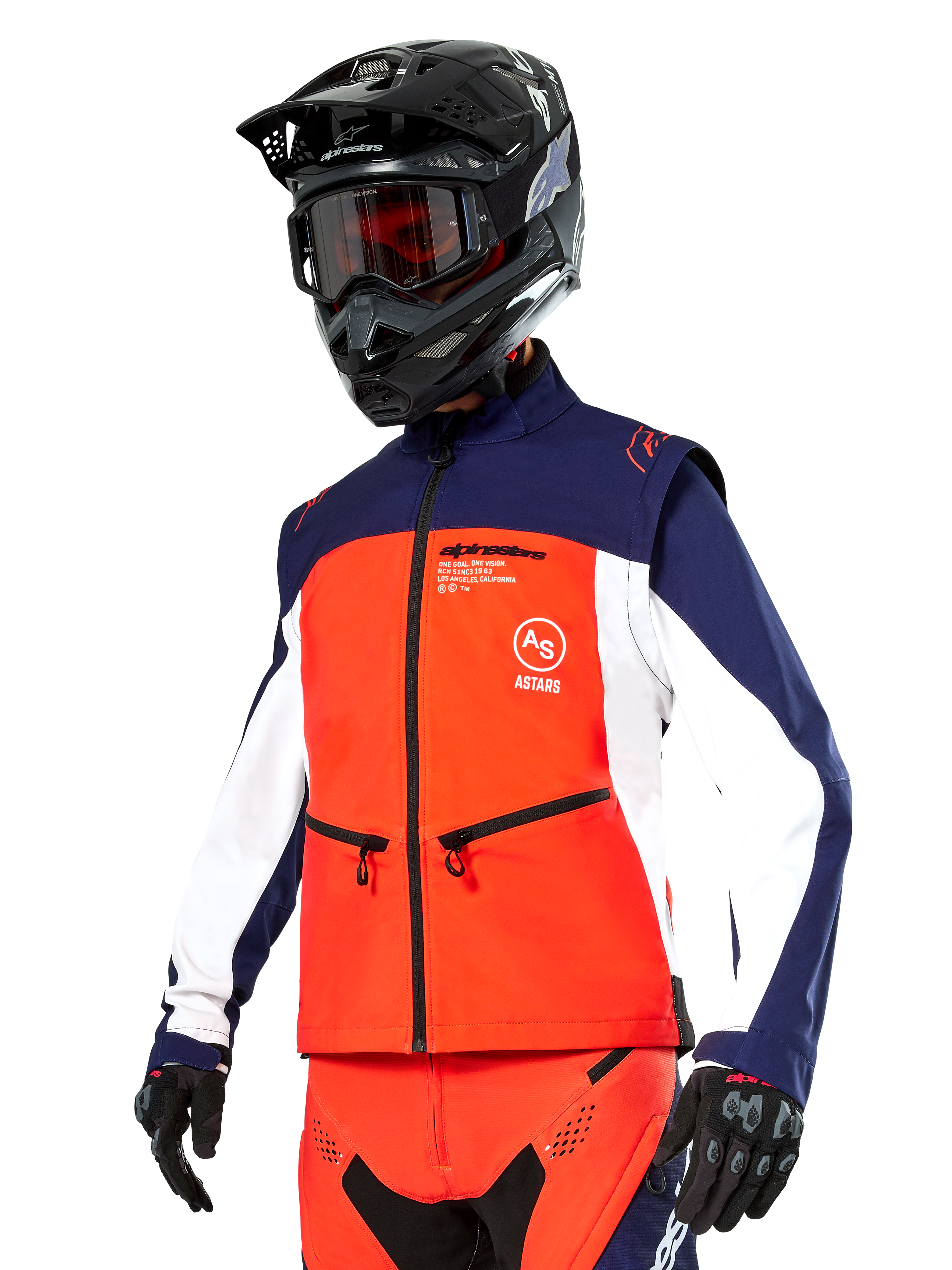 Veste Softshell Alpinestars Lite-Dura en bleu marine, orange vif et blanc, équipement de motocross, portée avec un casque et des lunettes noirs, des gants de protection et un pantalon de course assorti