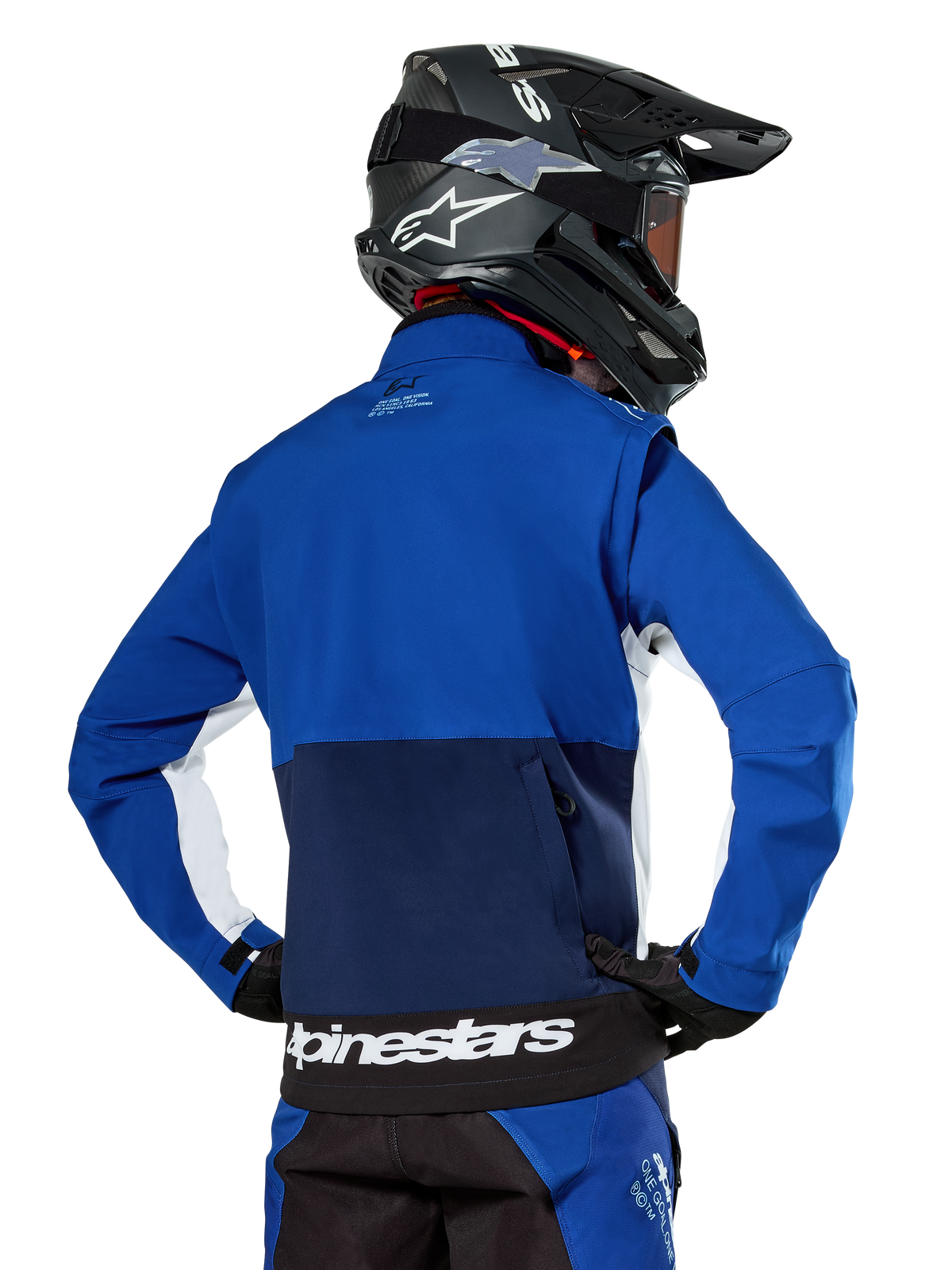 Veste Softshell Alpinestars Lite-Dura en bleu marine, bleu vif et blanc, vue arrière montrant la grande poche arrière zippée pour le rangement des manches, accents latéraux blancs et marquage Alpinestars sur l'ourlet, portée avec un casque de motocross noir et un équipement assorti.