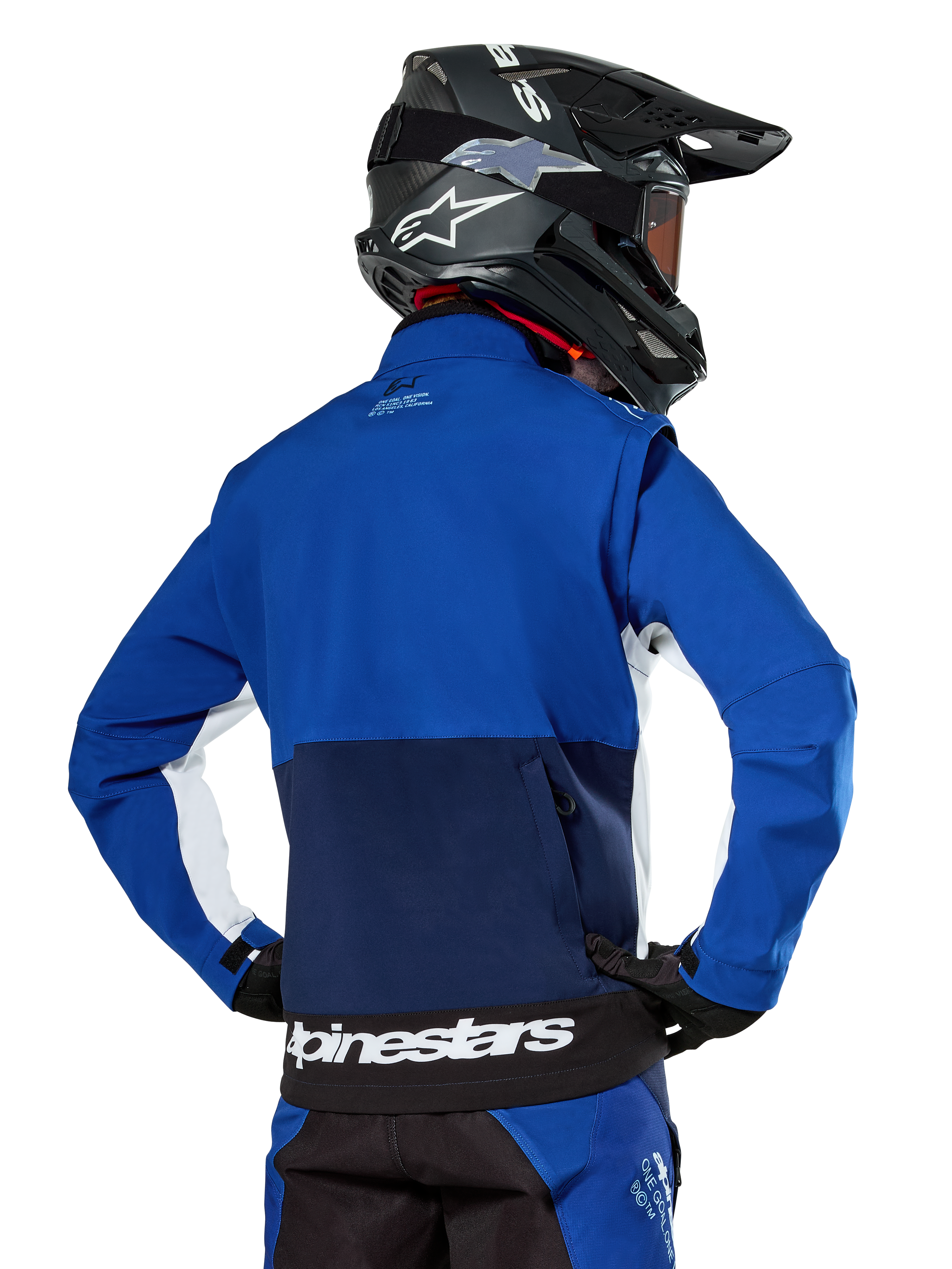 Alpinestars Lite-Dura Softshell Jacket in Navy, Bright Blue, and White, rear view showing the large zippered back pocket for sleeve storage, white side accents, and Alpinestars branding on the hem, worn with a black motocross helmet and matching gear.