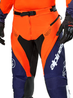 Pantalon Alpinestars Pro-Dura, pantalon de moto enduro, bleu marine, orange vif et blanc, vue détaillée de la taille avec boucle magnétique SX2 et ouvertures d'aération zippées sur les cuisses, matériau ripstop durable avec marquage distinctif, porté avec l'équipement assorti