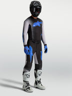 Pantalons Supertech Hiten AFD Alpinestars, pantalons de motocross pour homme, noirs et bleus, coupe athlétique performance, portés sur mannequin avec maillot assorti et bottes de moto blanches.