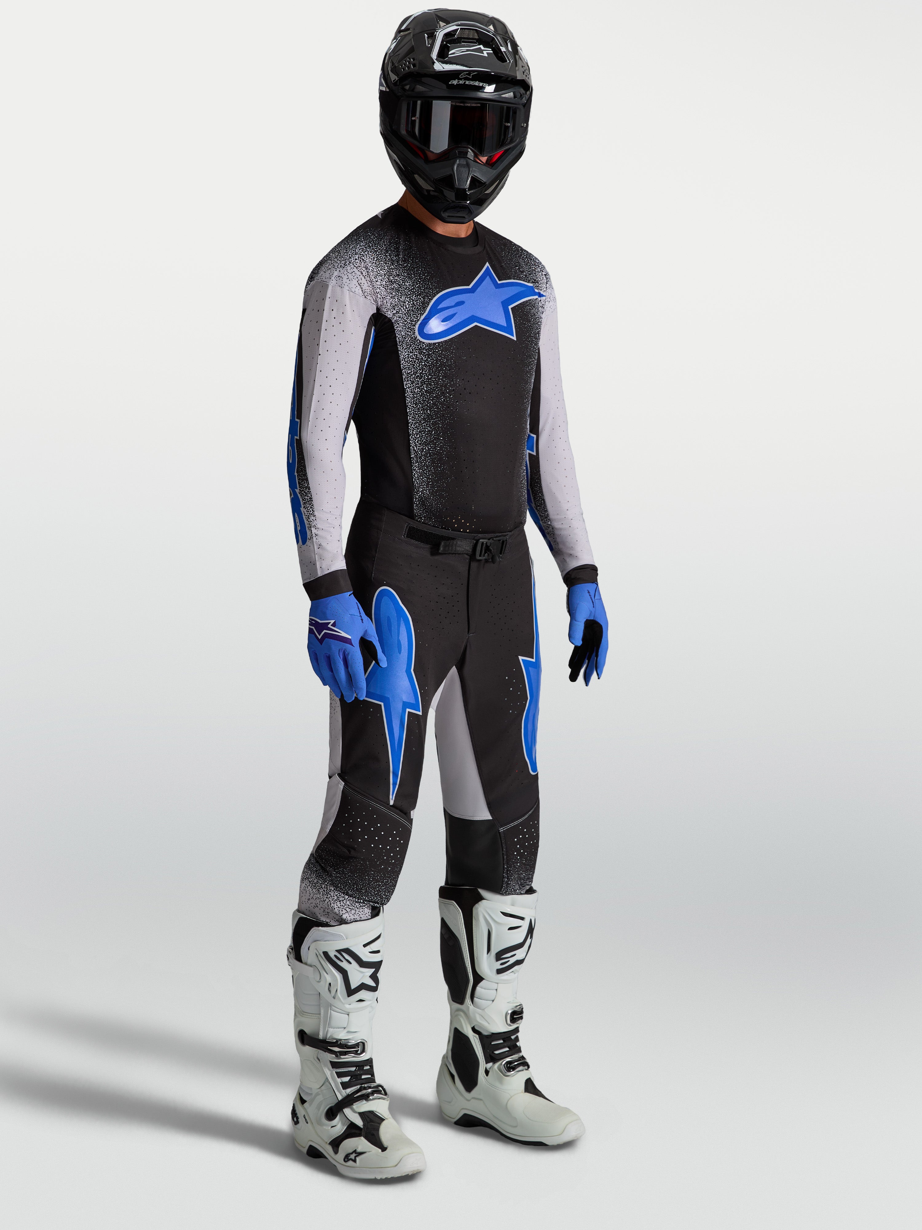 Pantalons Supertech Hiten AFD Alpinestars, pantalons de motocross pour homme, noirs et bleus, coupe athlétique performance, portés sur mannequin avec maillot assorti et bottes de moto blanches.