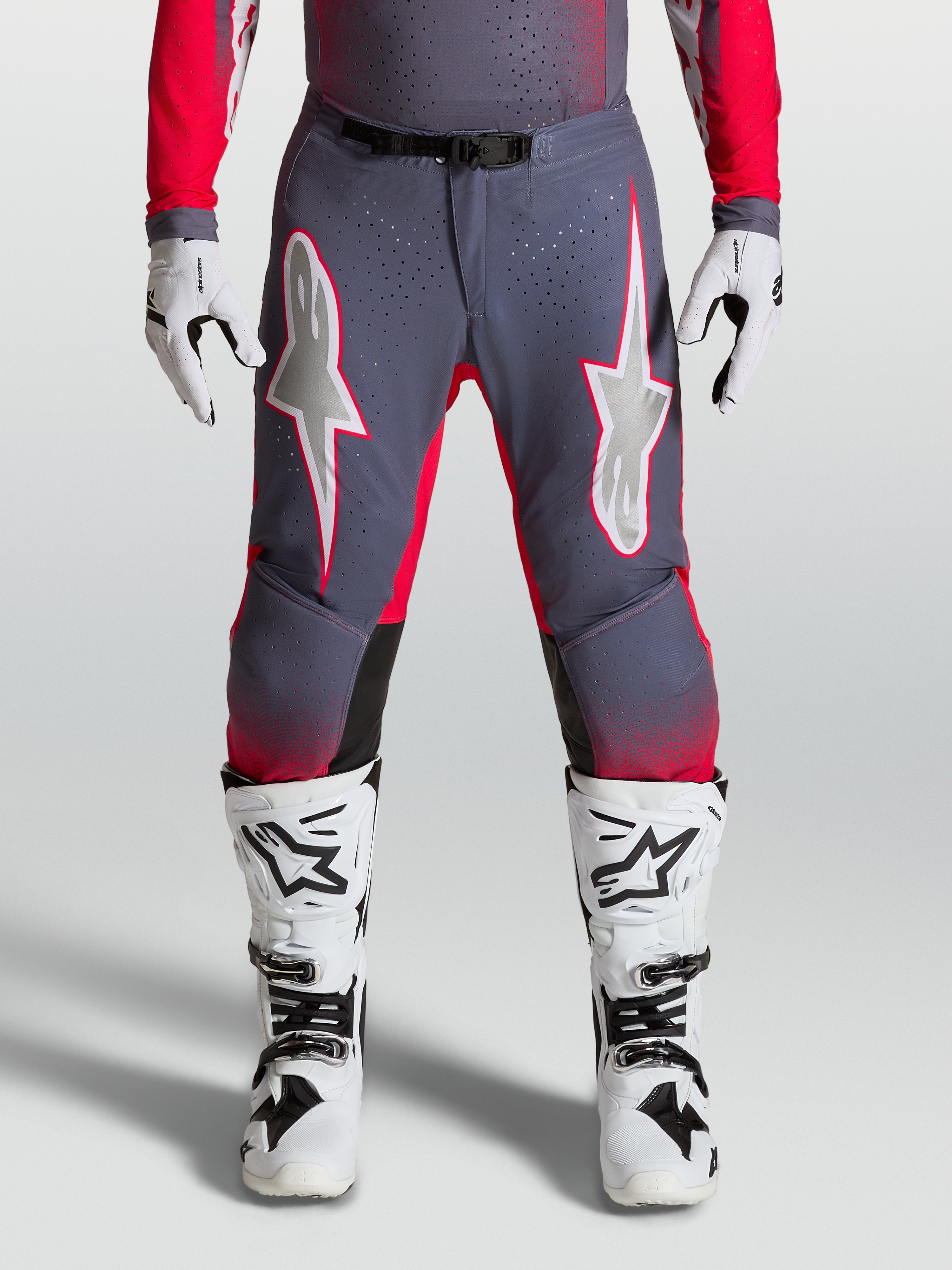 Alpinestars Supertech Hiten AFD, pantalon de motocross haute performance, gris et rouge fluo avec graphismes étoilés lilas clair, empiècements stretch perforés, porté avec des bottes blanches