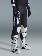 Pantalon Alpinestars 2026 Racer Air Riway, pantalon de motocross noir et blanc avec de grands graphismes de logo blancs, empiècements en cuir beige à l'intérieur du genou, porté avec des bottes tout-terrain blanches et noires et des gants noirs dans un décor de studio.