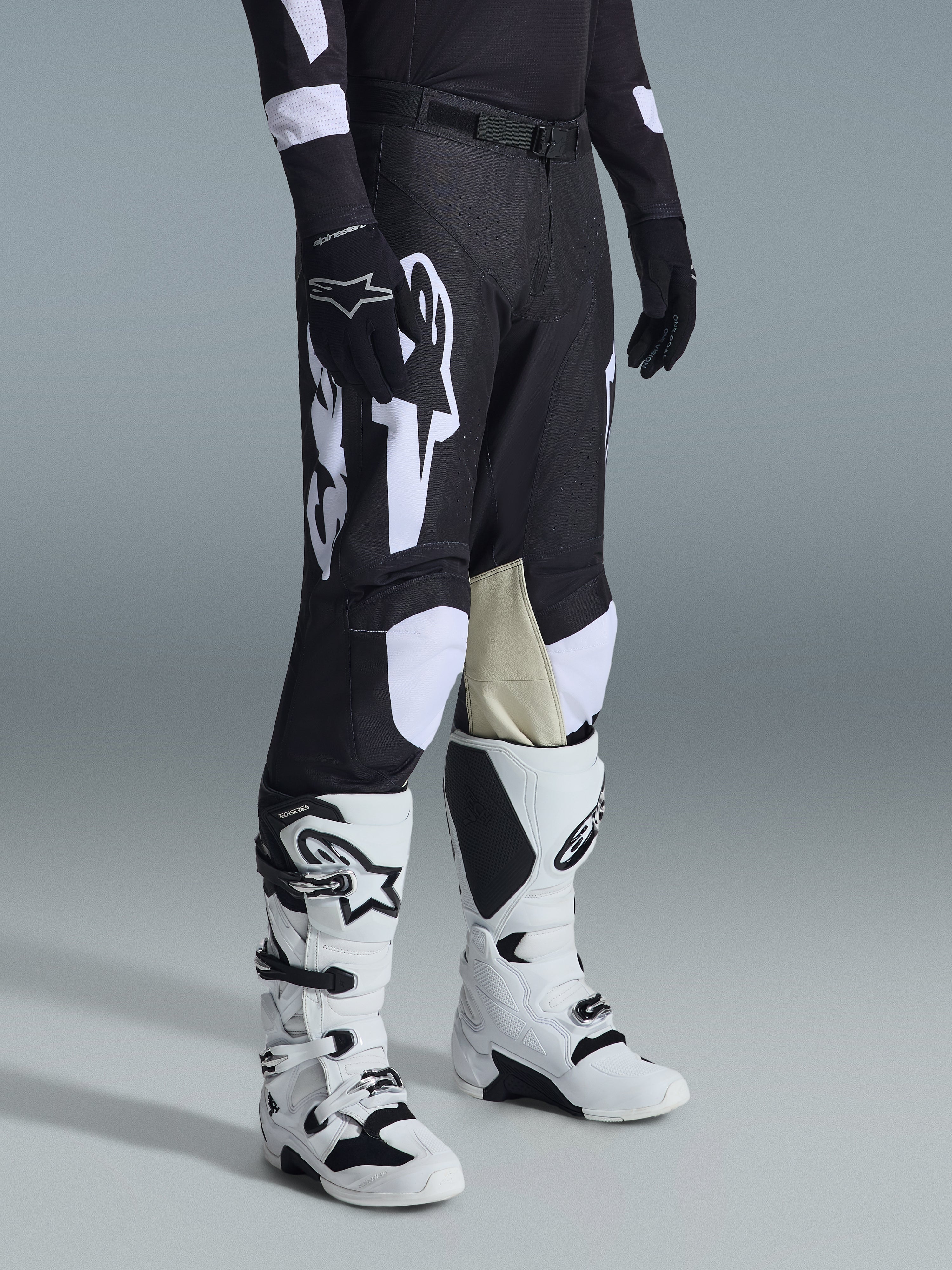 Pantalon Alpinestars 2026 Racer Air Riway, pantalon de motocross noir et blanc avec de grands graphismes de logo blancs, empiècements en cuir beige à l'intérieur du genou, porté avec des bottes tout-terrain blanches et noires et des gants noirs dans un décor de studio.