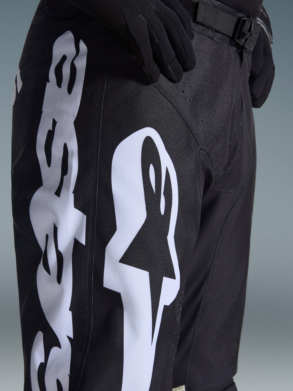 Pantalon Alpinestars Racer Air Riway 2026, pantalon de motocross noir avec un logo Alpinestars blanc sur la cuisse, gros plan montrant le tissu technique et la sangle de réglage à la taille, porté avec des gants noirs pour le pilotage tout-terrain