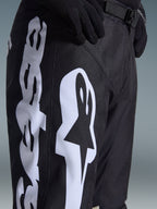 Pantalon Alpinestars Racer Air Riway 2026, pantalon de motocross noir avec un logo Alpinestars blanc sur la cuisse, gros plan montrant le tissu technique et la sangle de réglage à la taille, porté avec des gants noirs pour le pilotage tout-terrain