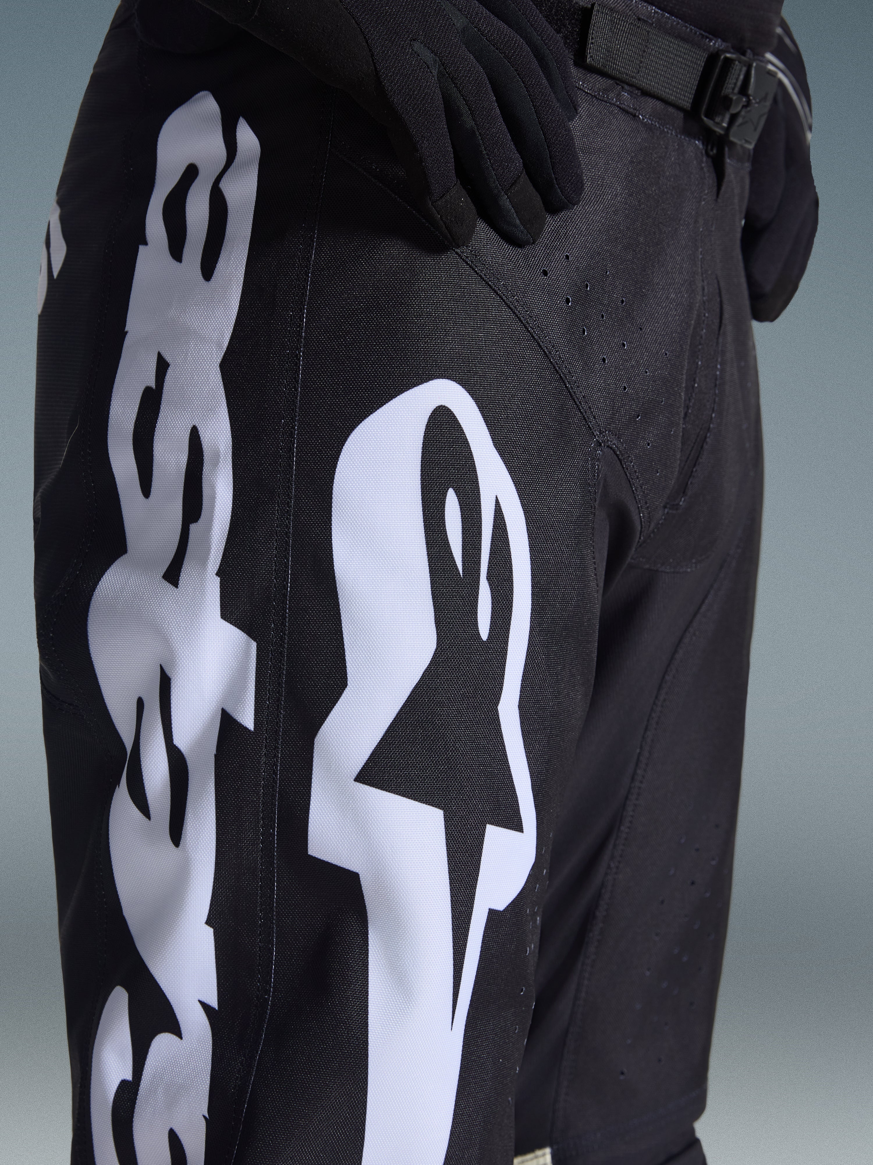 Pantalon Alpinestars Racer Air Riway 2026, pantalon de motocross noir avec un logo Alpinestars blanc sur la cuisse, gros plan montrant le tissu technique et la sangle de réglage à la taille, porté avec des gants noirs pour le pilotage tout-terrain