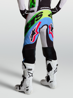 Alpinestars Pantalon Supertech Dyna, pantalon de motocross pour homme, lilas clair et vert fluo, porté par un pilote avec des bottes blanches, tissu rip-stop perforé et genoux en cuir de chèvre