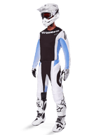Pilote de motocross portant le pantalon Alpinestars Techstar Melt 2025, tenue de motocross blanche et noire avec des accents bleu clair, maillot et gants coordonnés, casque et lunettes blancs, bottes de course blanches et noires, équipement tout-terrain professionnel.
