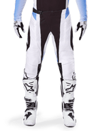Alpinestars Techstar Melt Pants 2025, pantalon de motocross, blanc et noir avec des accents bleu clair, porté avec des bottes et des gants blancs et noirs assortis, doté de perforations découpées au laser et d'un design de course haute performance.