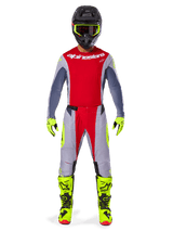 Pilote de motocross portant le pantalon Alpinestars Techstar Melt 2025 rouge vif et gris, avec des empiècements rouge éclatant et des accents jaune fluo, maillot assorti, casque noir et lunettes jaune fluo, bottes et gants jaune vif, tenue de course professionnelle coordonnée