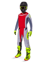 Pilote de motocross portant une tenue de course complète Alpinestars, maillot rouge et gris, pantalon Techstar Melt 2025 rouge vif et gris avec accents jaune fluo, bottes et gants jaune fluo, casque noir avec lunettes jaune fluo, tenue coordonnée pour la performance tout-terrain