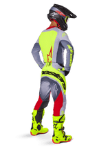 Pilote de motocross vu de trois-quarts arrière portant le pantalon Alpinestars Techstar Melt 2025 rouge vif et gris, avec un logo jaune fluo imposant sur l'assise, un maillot assorti gris et jaune fluo, des bottes jaune fluo et un casque noir avec lunettes fluo, tenue de course tout-terrain professionnelle.