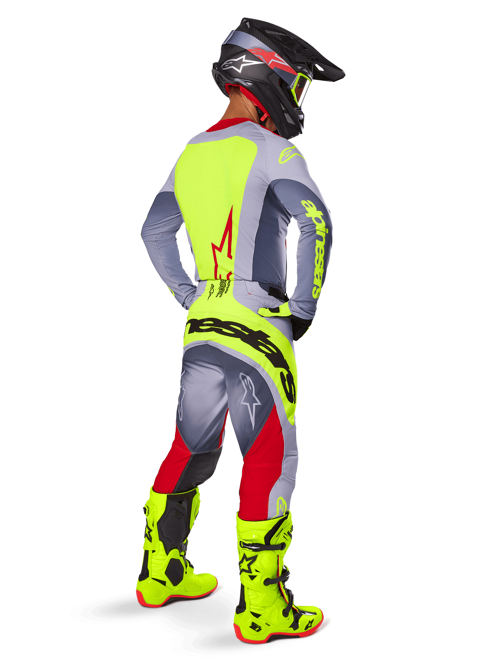 Pilote de motocross vu de trois-quarts arrière portant le pantalon Alpinestars Techstar Melt 2025 rouge vif et gris, avec un logo jaune fluo imposant sur l'assise, un maillot assorti gris et jaune fluo, des bottes jaune fluo et un casque noir avec lunettes fluo, tenue de course tout-terrain professionnelle.