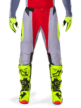 Alpinestars Techstar Melt Pants 2025, pantalon de motocross, rouge et gris avec accents jaune fluo, perforations découpées au laser pour la ventilation, porté avec des gants et des bottes jaune fluo, équipement haute performance pour la course tout-terrain