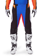 Pantalon de motocross Alpinestars 2025 Techstar Melt, orange et bleu, porté avec un maillot assorti et des bottes blanches, doté de panneaux noirs avec des accents bleu et orange vifs, perforé pour la ventilation, coupe athlétique de course pour la performance tout-terrain.