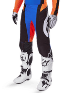 Pantalon de motocross Alpinestars Techstar Melt 2025, noir avec accents bleus et orange, porté avec des bottes blanches et un maillot assorti, doté d'un matériau de protection durable et d'un design de course sportif