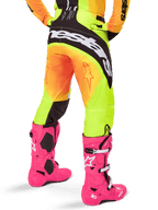 Vue arrière d'un pilote de motocross portant le pantalon Alpinestars Techstar Melt 2025, dégradé jaune et orange avec logo noir et blanc sur l'assise, associé à des bottes de motocross rose vif, équipement tout-terrain haute performance