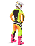 Pilote de motocross vu de dos portant l'ensemble Alpinestars Techstar Melt 2025, maillot et pantalon avec dégradé jaune et orange et empiècements noirs, casque blanc avec masque rouge, bottes rose vif, tenue de course coordonnée pour la performance tout-terrain