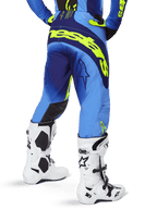 Alpinestars Pantalon Techstar Melt 2025, pantalon de motocross, bleu et jaune fluo, vue arrière montrant le large logo jaune sur l'assise et les cuisses, porté avec des bottes de motocross blanches et un maillot bleu assorti, coupe athlétique pour la conduite tout-terrain