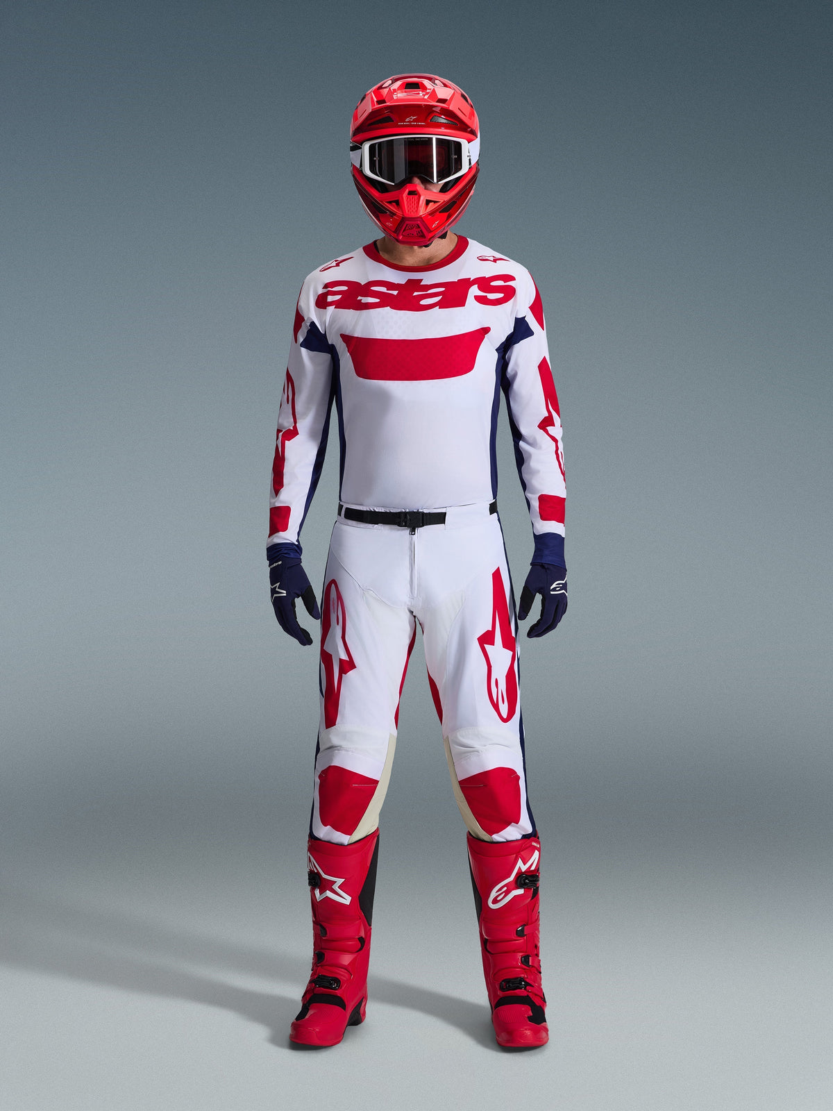Motocross rider wearing the Alpinestars 2026 Racer Riway Gear Set, White jersey with red "astars" branding and blue side panels, white pants with red graphics, red racing helmet and boots, navy blue gloves, complete coordinated motocross racing outfit