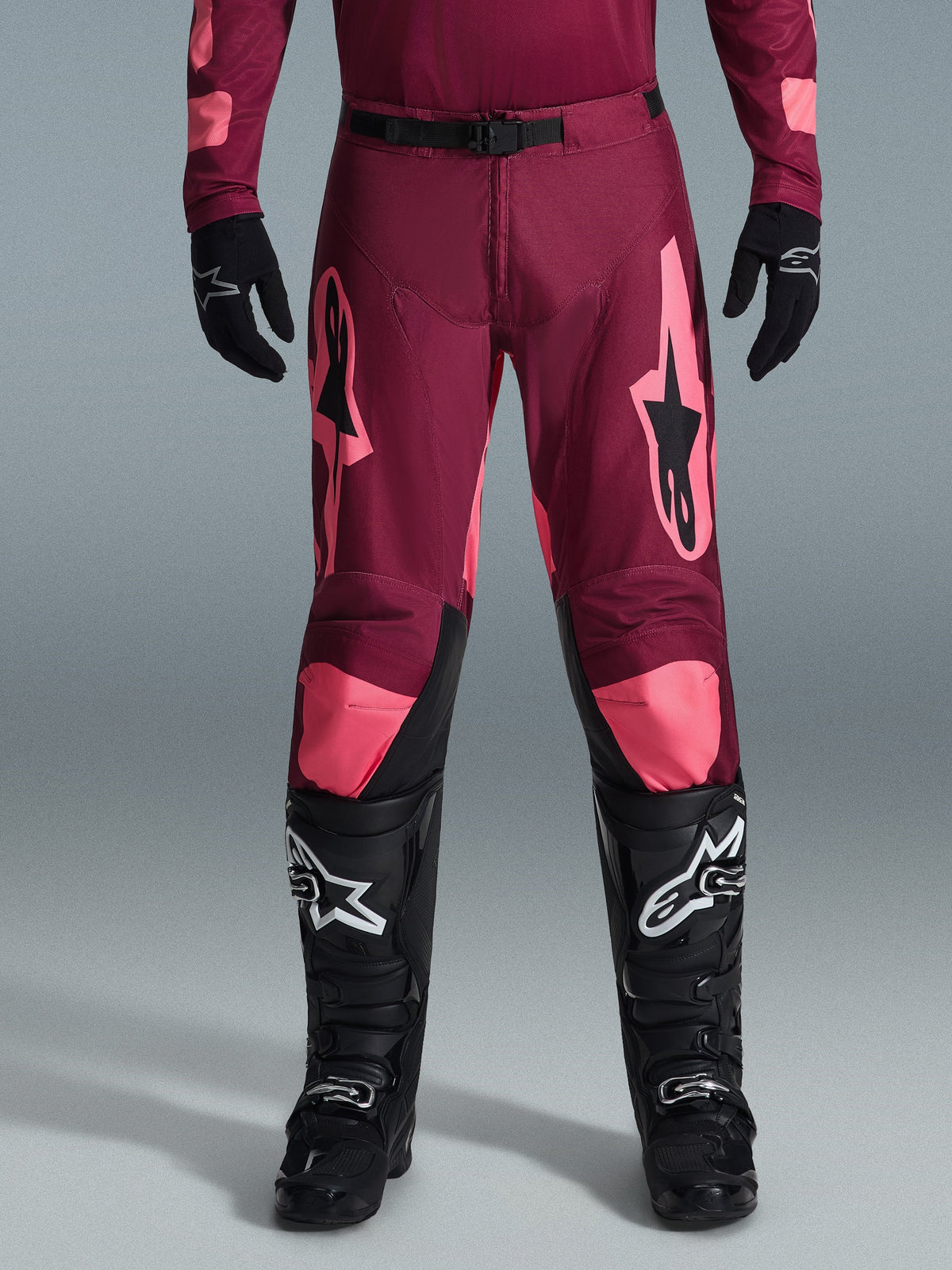 Pantalon Alpinestars Racer Riway 2026, pantalon de motocross, rouge corail foncé, porté avec des gants et des bottes noirs, avec de grands logos Alpinestars corail sur les cuisses, boucle noire à la taille et jambes pré-courbées pour la course tout-terrain