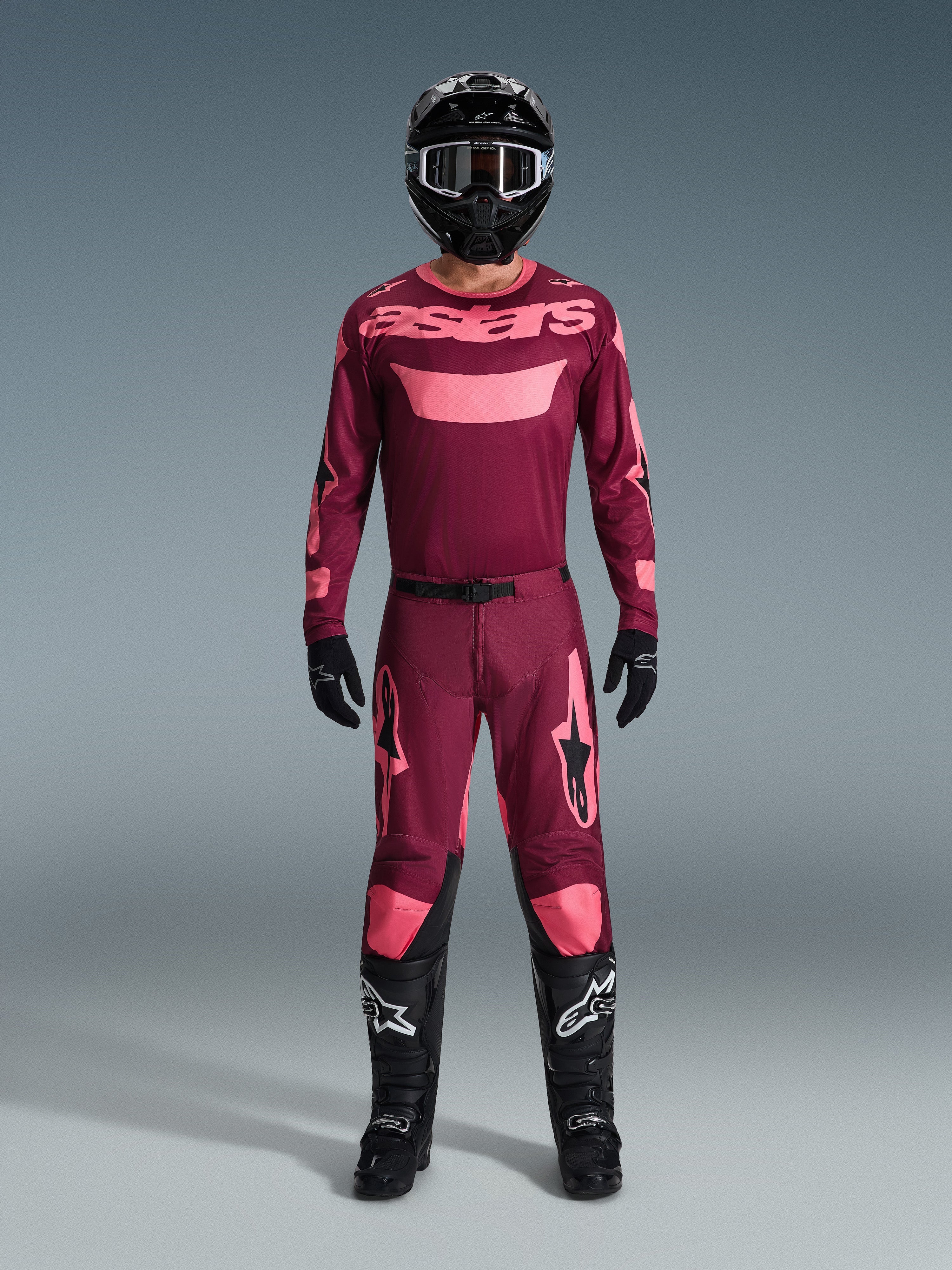 Pilote de motocross portant l'ensemble Alpinestars 2026 Racer Riway, maillot bordeaux avec marquage 'astars' rose, pantalon bordeaux assorti avec accents roses aux genoux et aux jambes, casque noir, gants noirs et bottes noires, tenue de performance complète pour la course tout-terrain