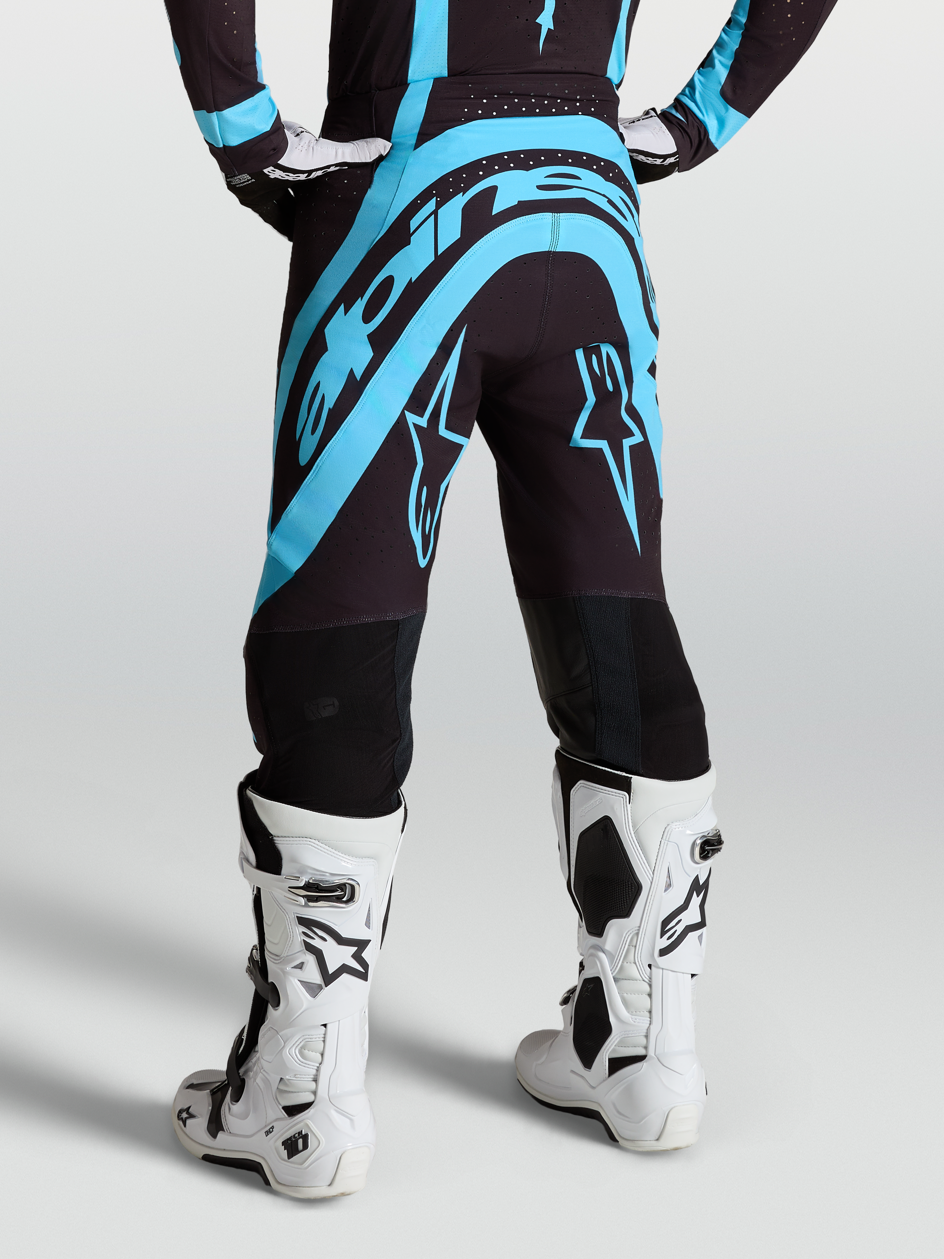 Pantalon Supertech Vesta Alpinestars, équipement de motocross noir et turquoise, vue arrière sur mannequin avec bottes blanches.