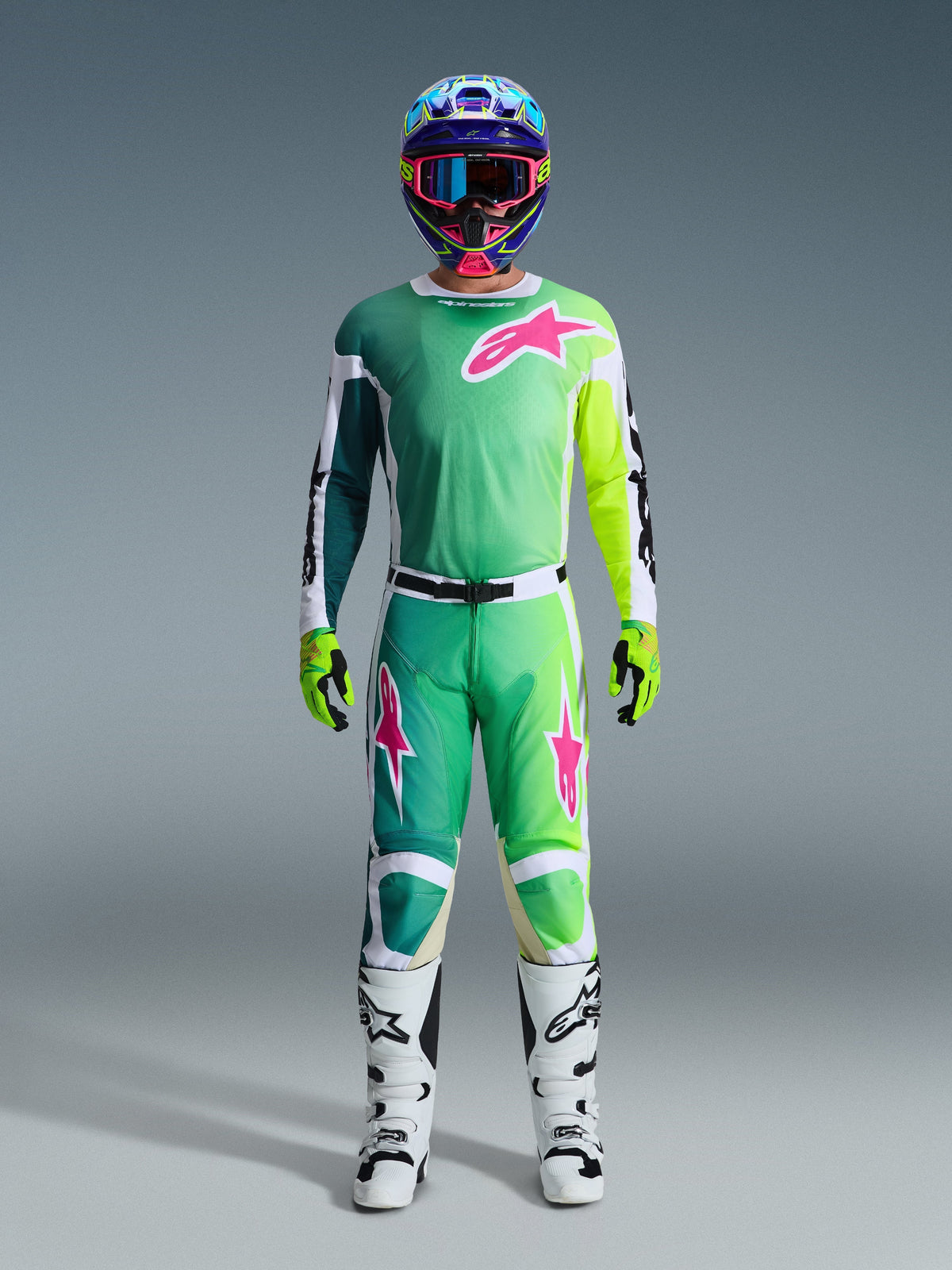 Alpinestars 2026 Racer Portl Gear Set, pilote de motocross portant une tenue de course complète, maillot et pantalon verts avec graphismes roses et blancs, casque bleu multicolore, bottes blanches et noires et gants verts, tenue coordonnée pour la course tout-terrain