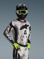 Motocross rider wearing the Alpinestars 2026 Racer Lahnd Jersey and gear set in Sand Camo, light gray camouflage pattern with black logo branding, black helmet with white and neon yellow goggles, bright neon yellow gloves, professional off-road racing apparel