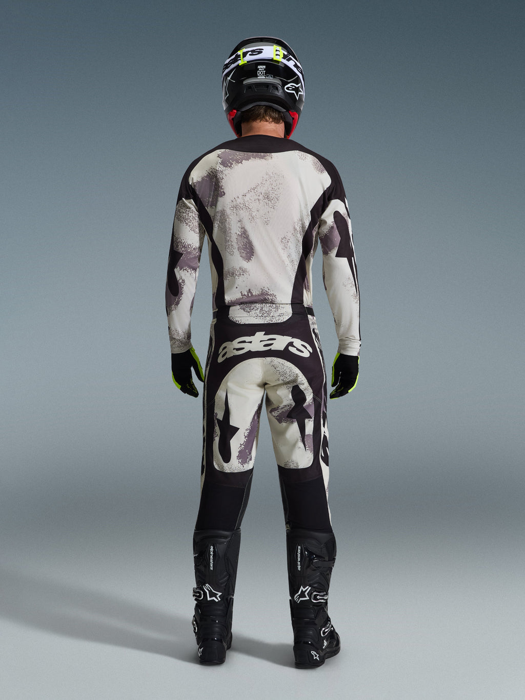 Motocross rider wearing complete Alpinestars 2026 Racer Lahnd Gear Set, viewed from behind, Sand Camo jersey and pants with light gray and black abstract pattern, black helmet, black and neon yellow gloves, black boots, featuring 'astars' branding on the rear of the pants
