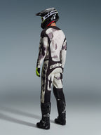 Motocross rider wearing complete racing gear, 2026 Racer Lahnd Jersey and matching pants in Sand Camo, featuring a gray and black camouflage pattern with bold Alpinestars logos, black helmet, black boots, and neon yellow gloves, rear profile view.