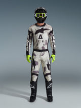Motocross rider wearing complete racing gear, Alpinestars 2026 Racer Lahnd Pants and matching jersey in Sand Camo gray, black helmet with neon yellow goggles, bright neon yellow gloves, and black boots, coordinated professional outfit for off-road racing