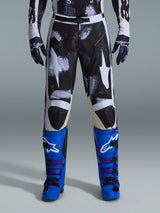 Alpinestars 2026 Racer Lahnd Pants, pantalon de motocross, Iron Camo, porté avec des gants de camouflage assortis et des bottes bleu vif et violet, arborant des graphismes d'étoiles blanches stylisées sur un motif camouflage sombre, fermeture technique par boucle magnétique, équipement de course tout-terrain professionnel.