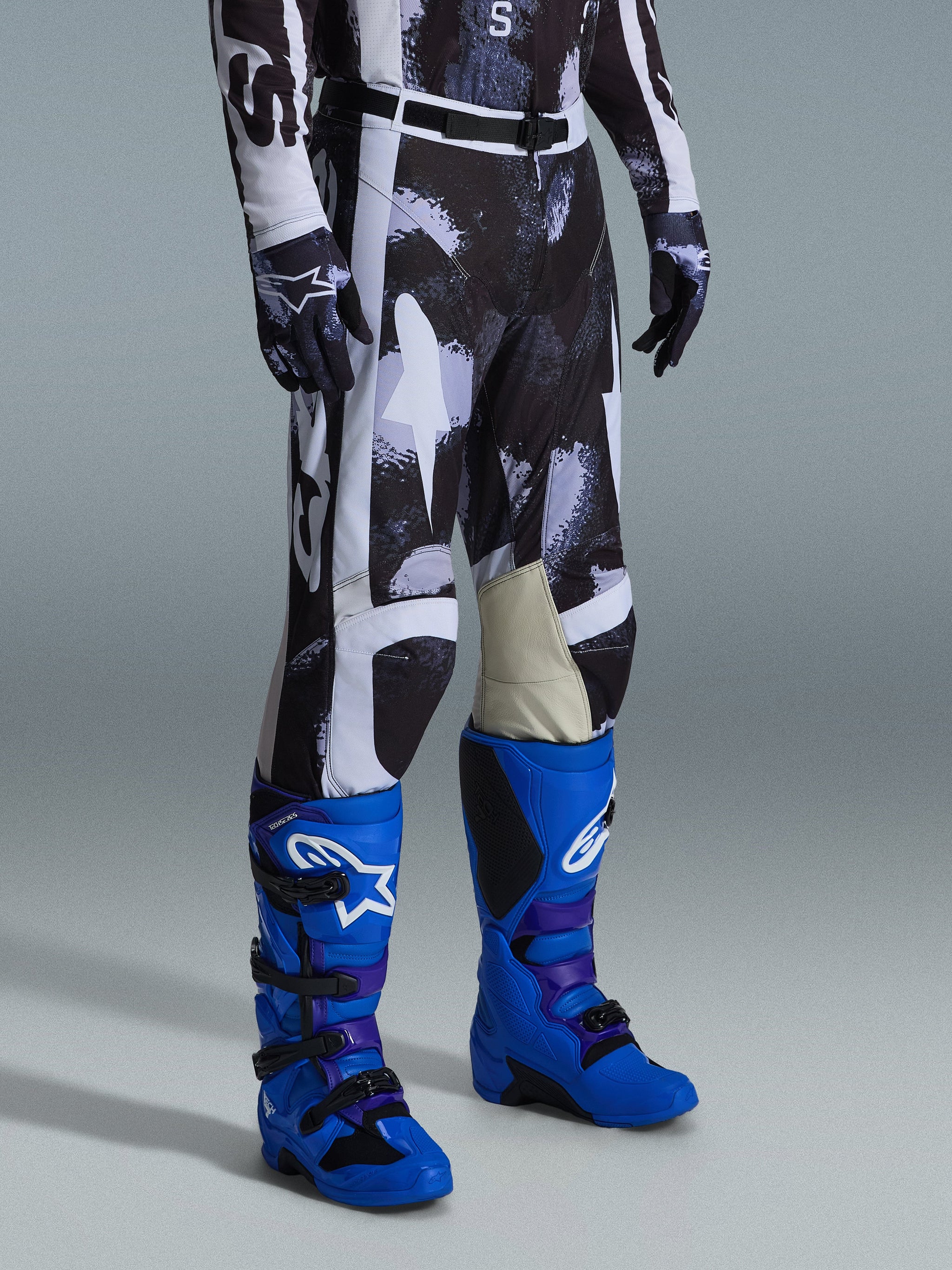 Pantalon Alpinestars Racer Lahnd 2026, pantalon de motocross, motif camouflage Iron Camo gris et noir, porté avec des bottes de course bleu vif et des gants assortis, doté de panneaux blancs et de genoux intérieurs en cuir de chèvre, équipement tout-terrain axé sur la performance