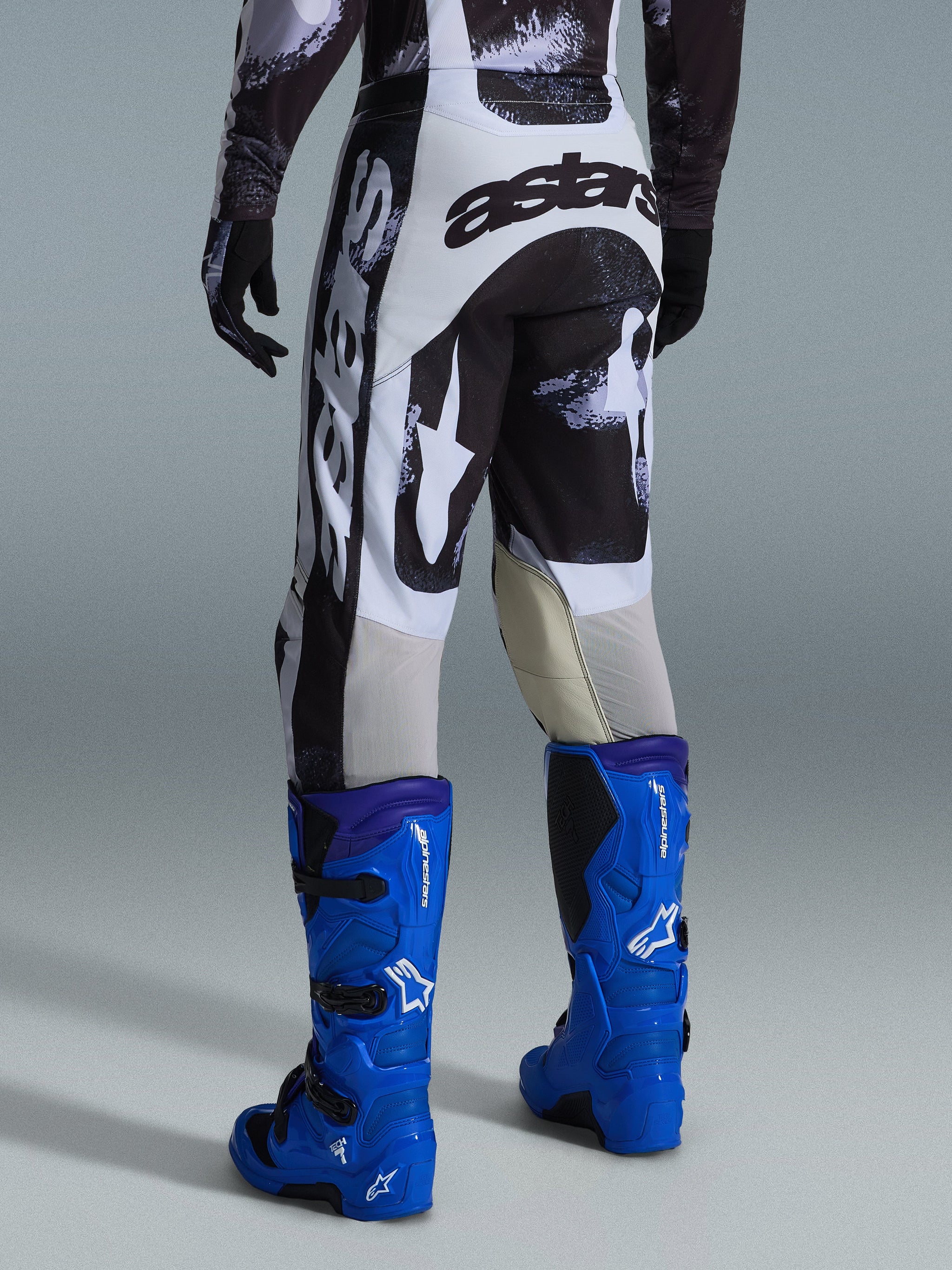 Pantalon Alpinestars Racer Lahnd 2026, pantalon de motocross, motif Iron Camo gris et noir, vue arrière montrant un large marquage astars blanc et noir, associé à des bottes de motocross bleu vif, équipement de course tout-terrain professionnel