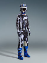 Pilote de motocross portant un équipement complet comprenant le pantalon Alpinestars Racer Lahnd 2026, motif Iron Camo et gris, maillot assorti, bottes de motocross bleu vif et violet, casque bleu et blanc, tenue de course tout-terrain professionnelle.