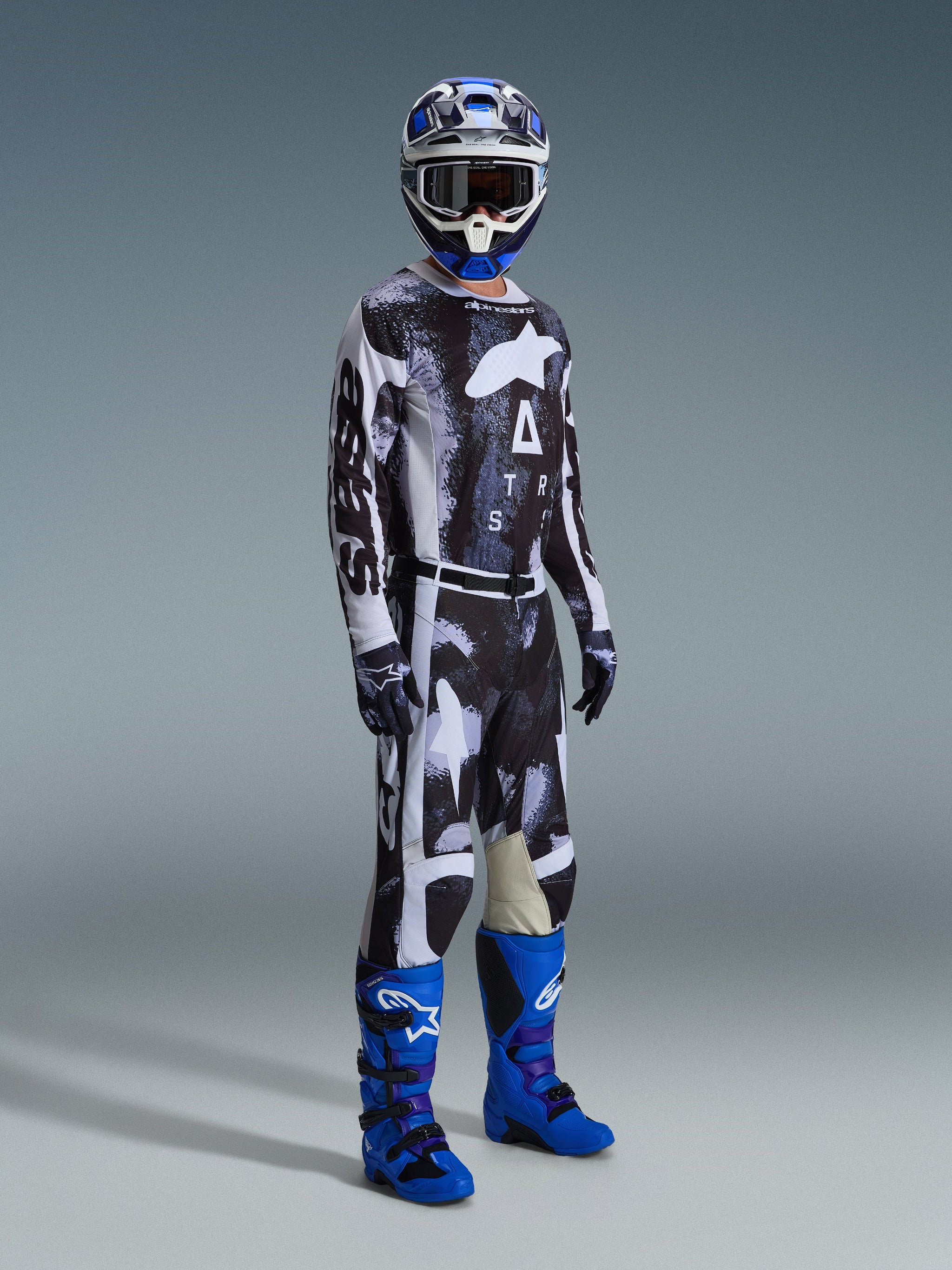 Pilote de motocross portant un équipement complet comprenant le pantalon Alpinestars Racer Lahnd 2026, motif Iron Camo et gris, maillot assorti, bottes de motocross bleu vif et violet, casque bleu et blanc, tenue de course tout-terrain professionnelle.