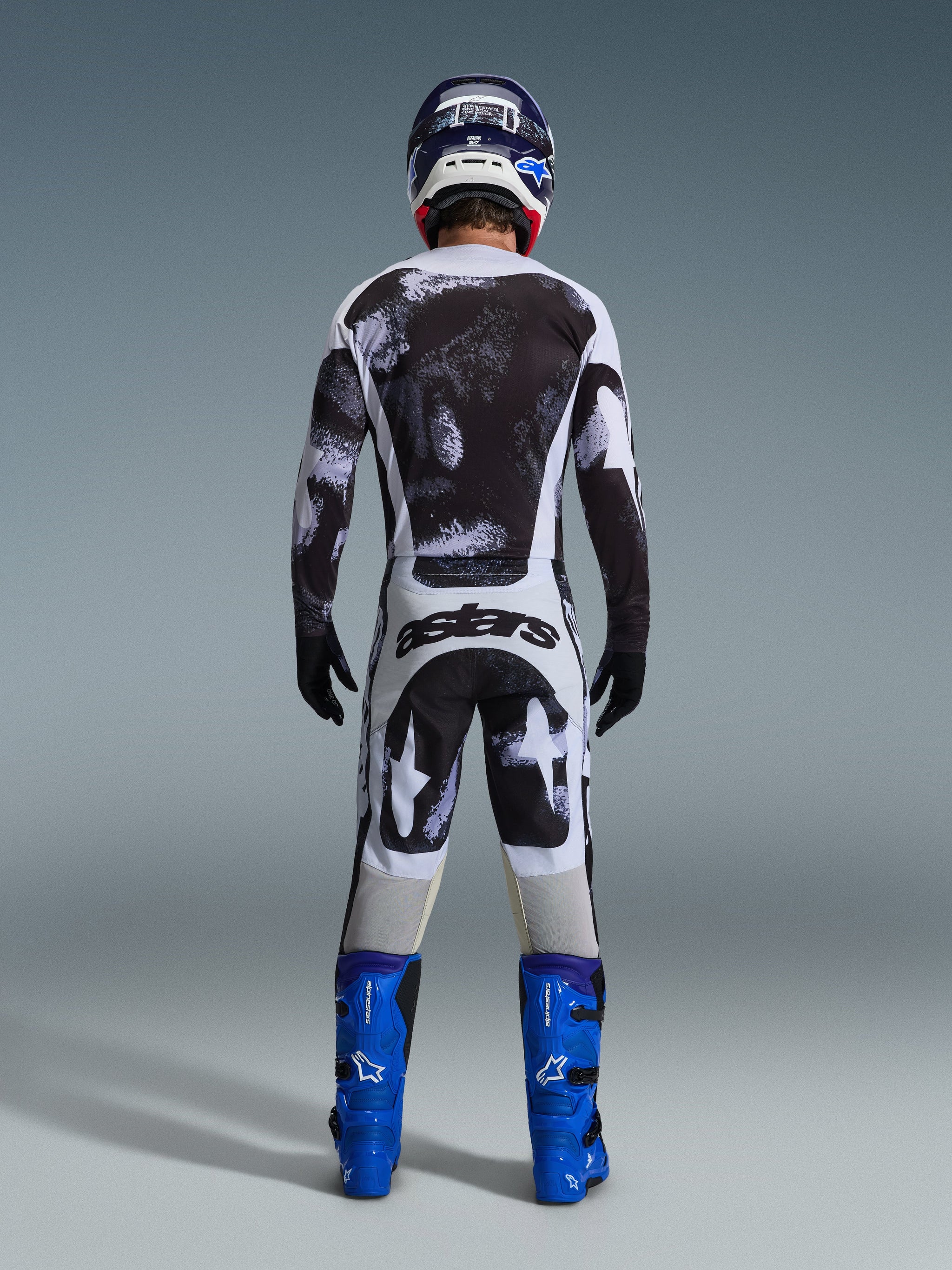 Pantalon Alpinestars 2026 Racer Lahnd, pantalon de motocross, motif Iron Camo gris et noir, vue arrière du pilote en équipement de course complet avec maillot assorti et bottes bleues, conçu pour la performance tout-terrain.