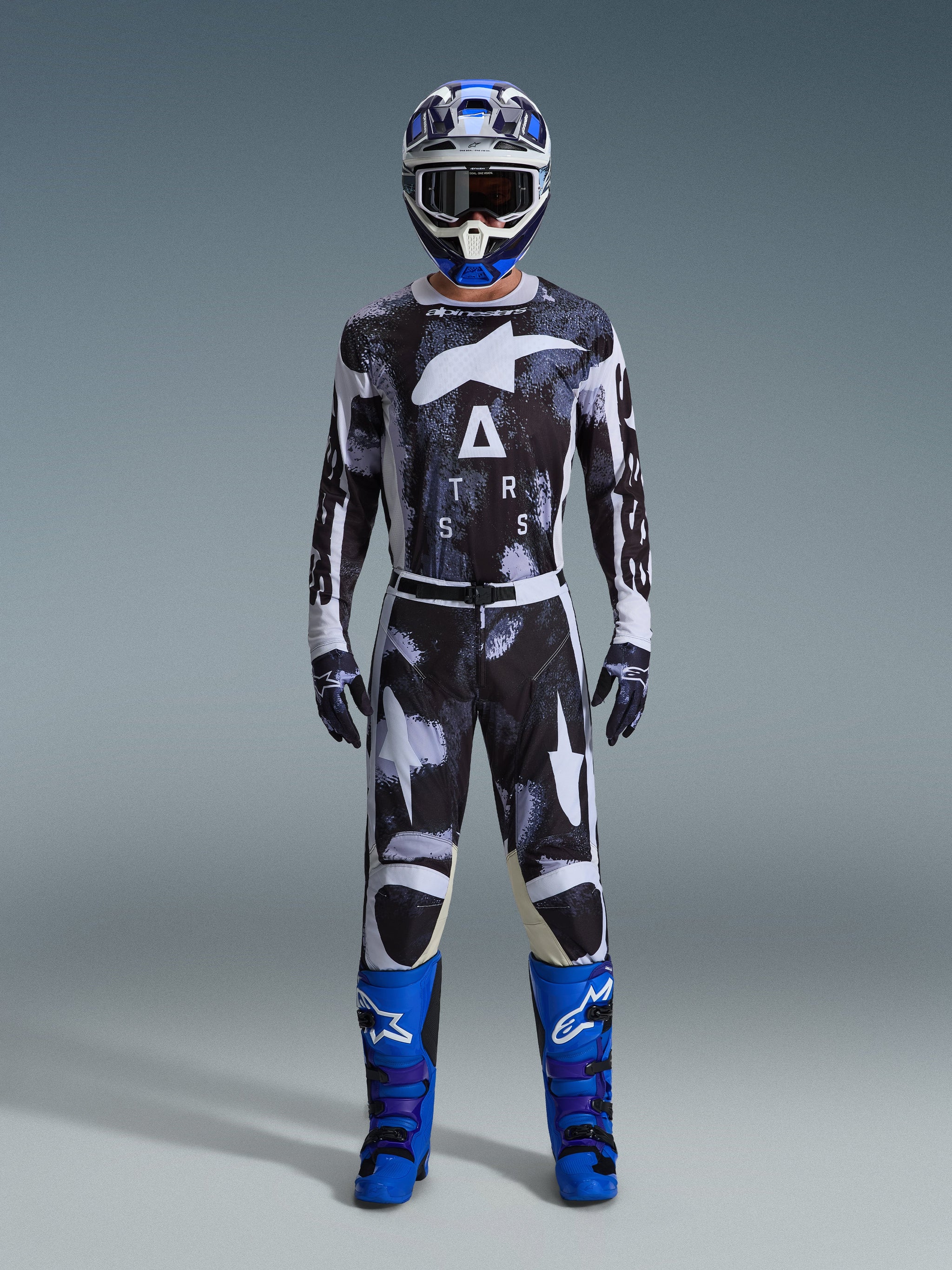 Pilote de motocross portant le pantalon Racer Lahnd 2026 d'Alpinestars en Iron Camo, motif camouflage gris et noir, maillot assorti, casque bleu et blanc avec lunettes, et bottes bleu vif, ensemble d'équipement de motocross coordonné.